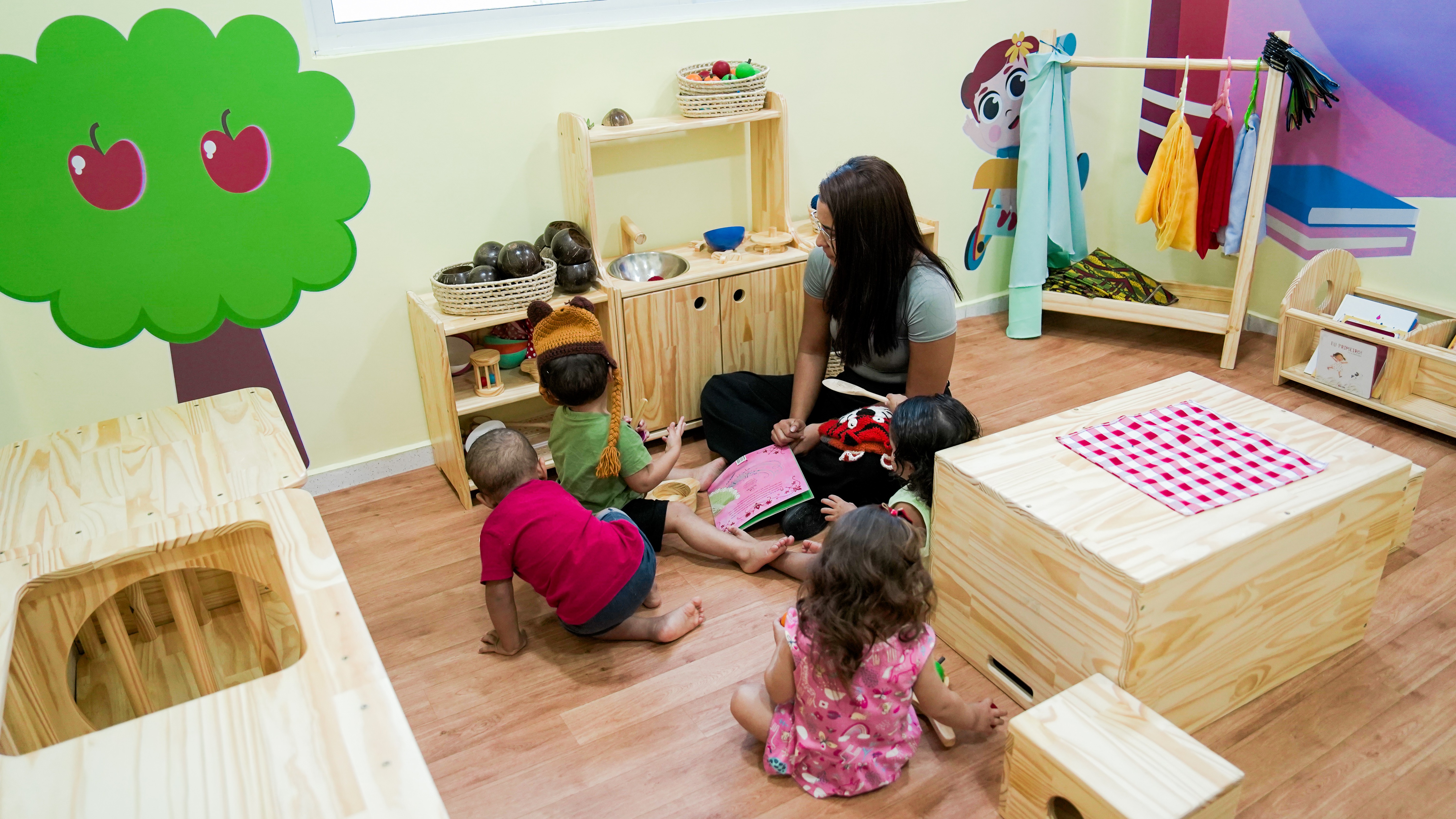 Três crianças de costas, sem aparecer o rosto, sentadas num chão de uma bebeteca ao lado de uma cuidadora; o espaço conta com brinquedos e mobiliário infantil